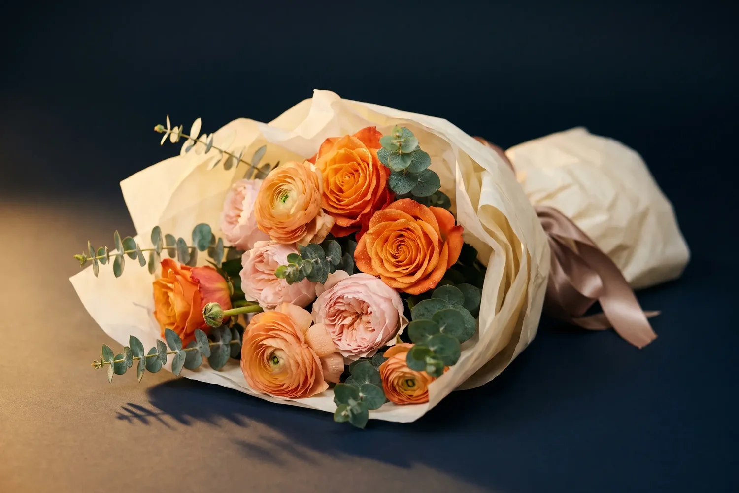 A hand-tied bouquet of orange roses, peach ranunculus and eucalyptus in cream paper
